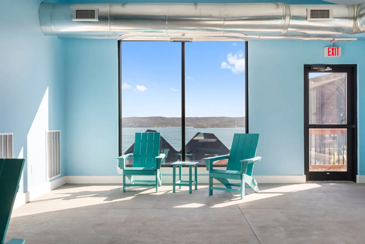 A cozy interior nook with two turquoise Adirondack chairs facing a large window overlooking a tranquil lake. A small table sits between the chairs, perfect for a coffee break or casual work session, with the calming water view in the background.