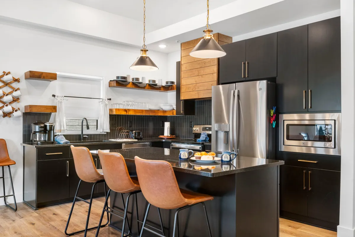 A warm, modern kitchen interior with a spacious island, bar stools, hanging lights, and wooden shelves. The area is inviting and set up for cooking, entertaining, or gathering after a day out.