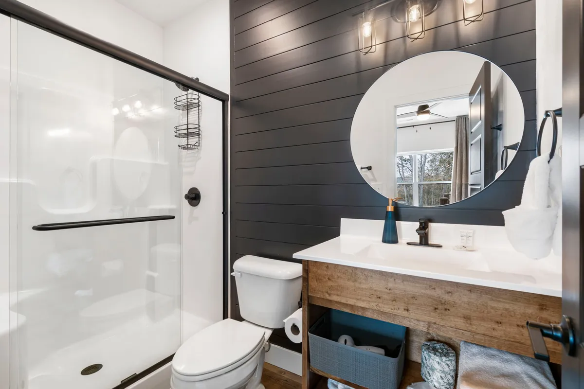 A stylish ensuite bathroom featuring a circular mirror, wooden vanity, sleek shower area, and a striking black accent wall. The fixtures are modern, and the scene feels fresh and contemporary, ideal for self-care routines.