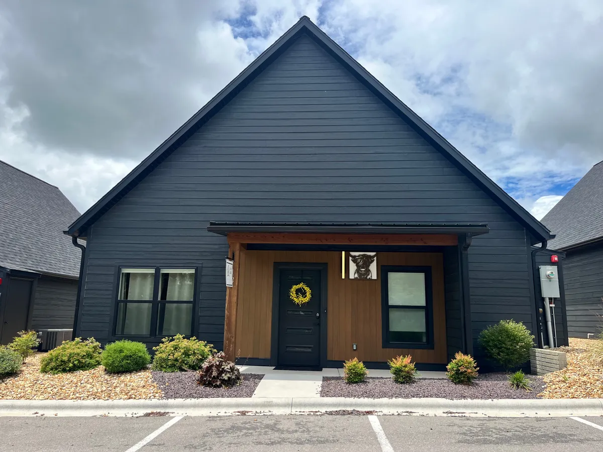 A charming exterior shot of a cozy cabin featuring dark wooden siding, light wood accents, and a welcoming yellow wreath on the door. The scene evokes the peacefulness of morning with the suggestion of sipping coffee on the porch, surrounded by nature.
