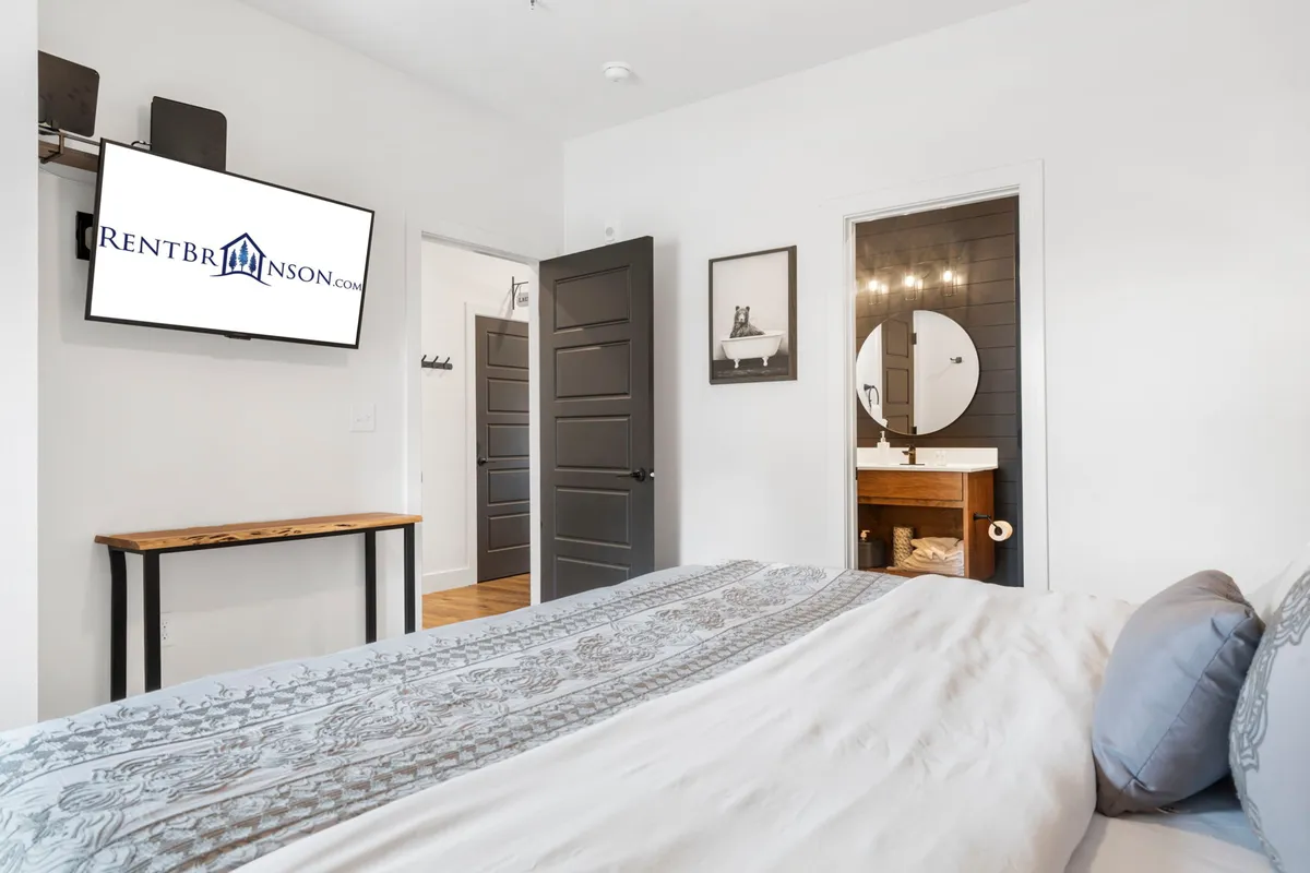 This image features a king bedroom with decorative throw pillows on the bed, a mounted television, a door leading to an ensuite bathroom highlighted by a circular mirror, and tasteful wall artwork. The ambiance is comfortable and contemporary, emphasizing relaxation and privacy.
