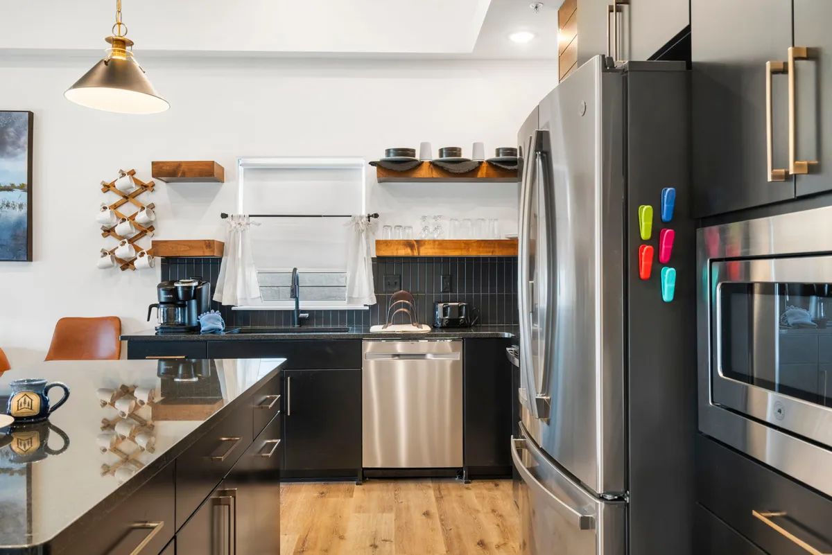 A bright, modern kitchen with a spacious island, stainless steel appliances, open wooden shelving, and colorful magnets on the refrigerator. The space is spotless and designed for both culinary creativity and social gatherings.