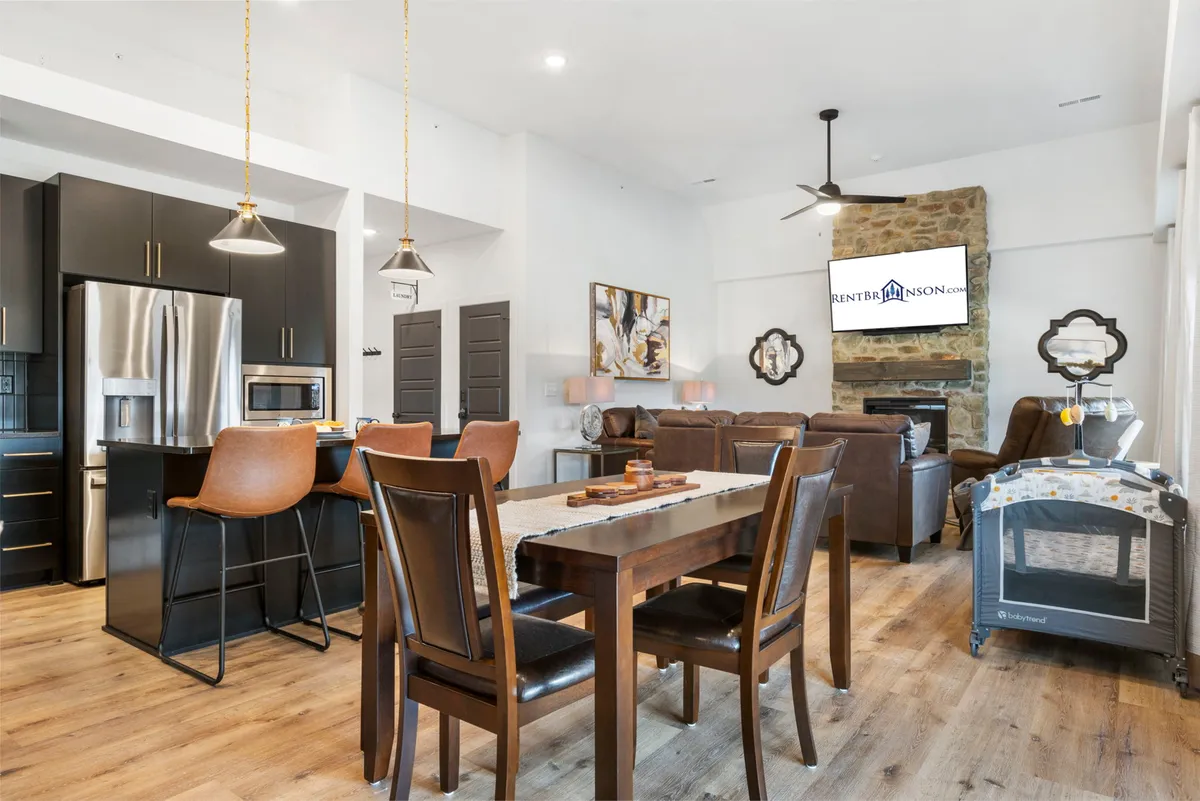An open-concept living area featuring a kitchen island, dining table, cozy living room seating, a stone fireplace, and a TV. The kitchen seamlessly flows into the living area, creating a unified and inviting space for cooking, dining, and relaxing.