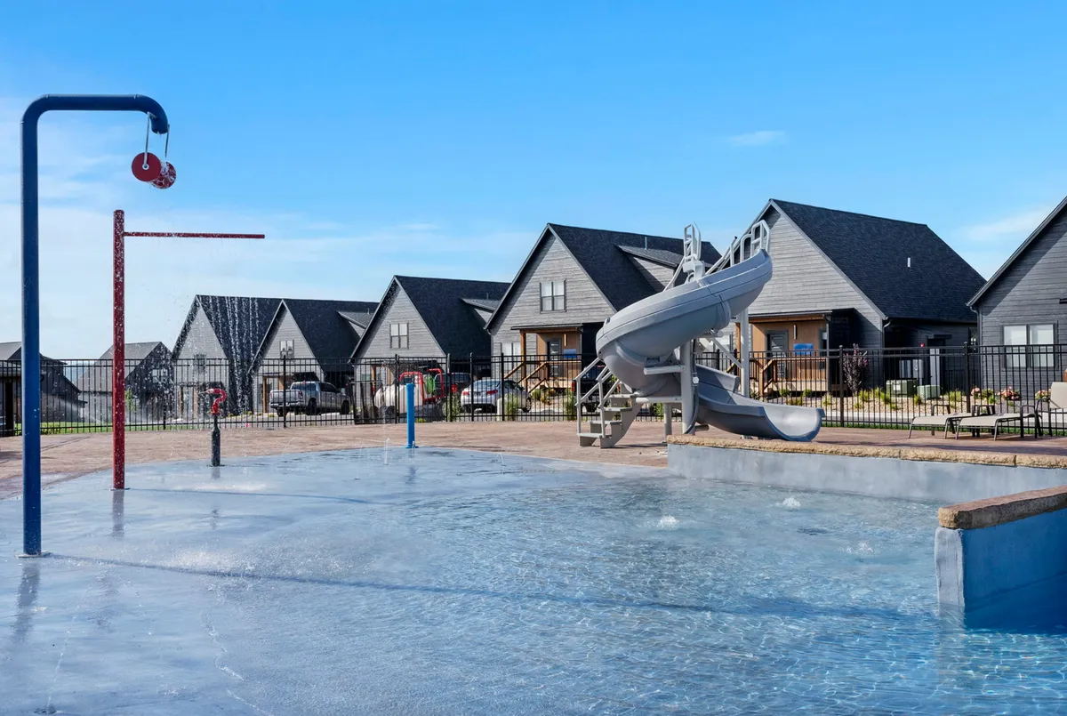 A vibrant exterior scene featuring a splash pad, water slide, and swimming pool surrounded by neighboring houses. Children and families are actively playing and splashing in the water, capturing lots of excitement and togetherness.